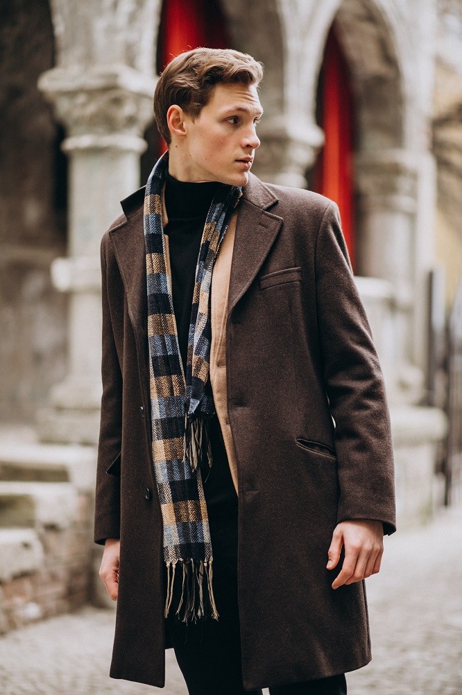A younger man is wearing a long brown jacket, blue scarf, and tan sweater