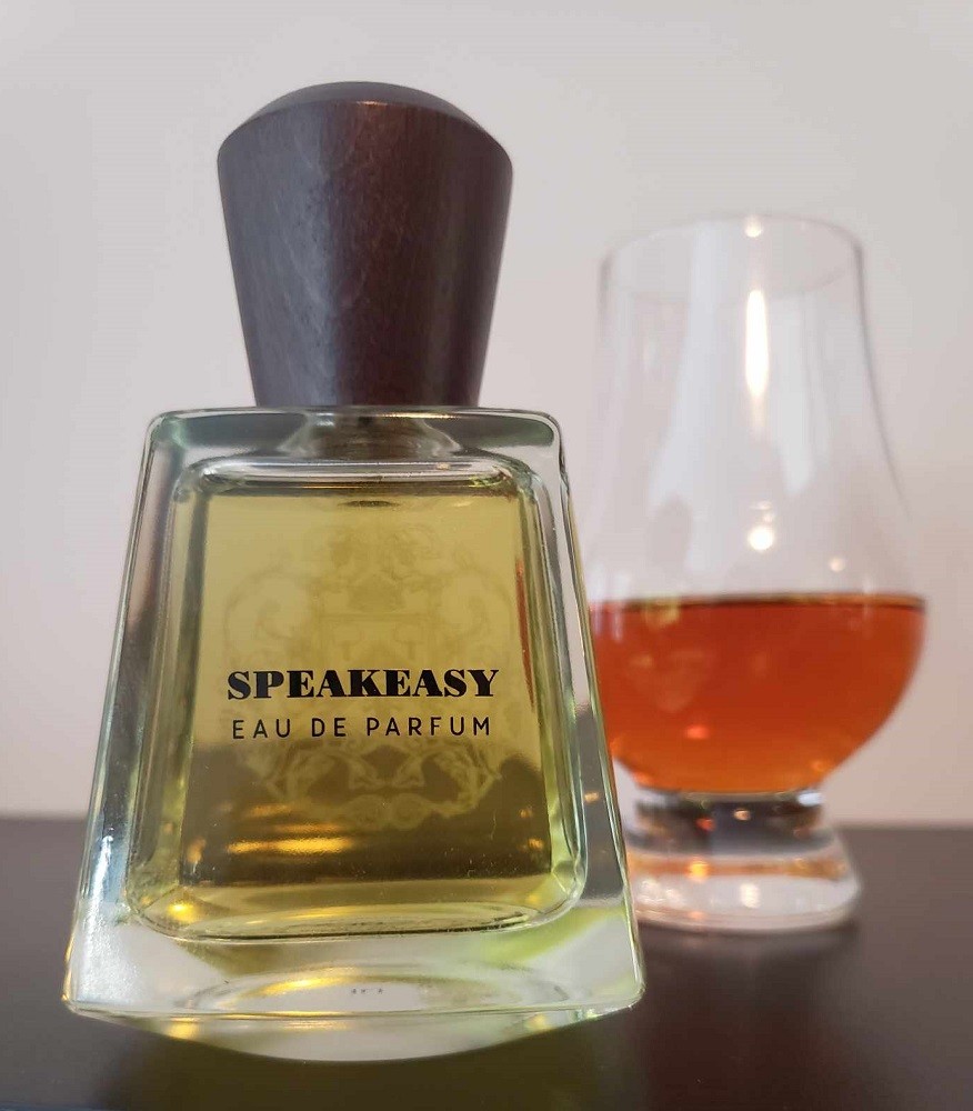 A bottle of Speakeasy cologne by Frapin sits in front of a glencarin glass of amber brown whiskey