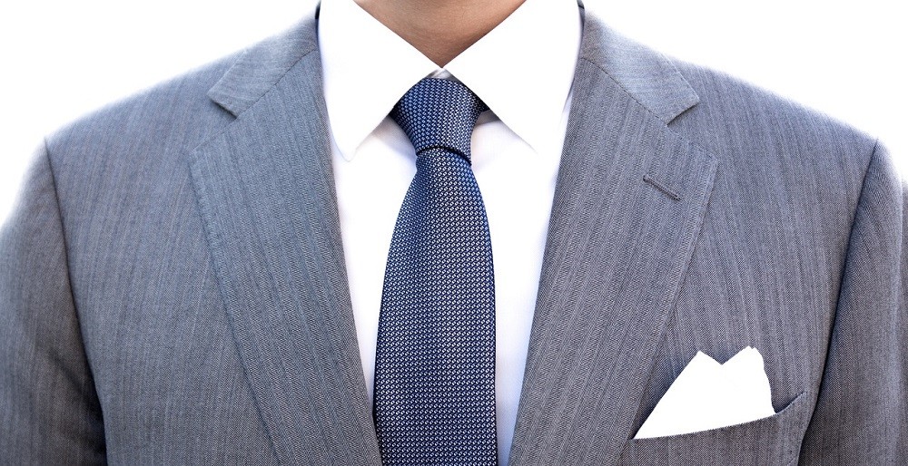 A man wearing a white dress shirt and light blue suit has a dark blue tie and white pocket square