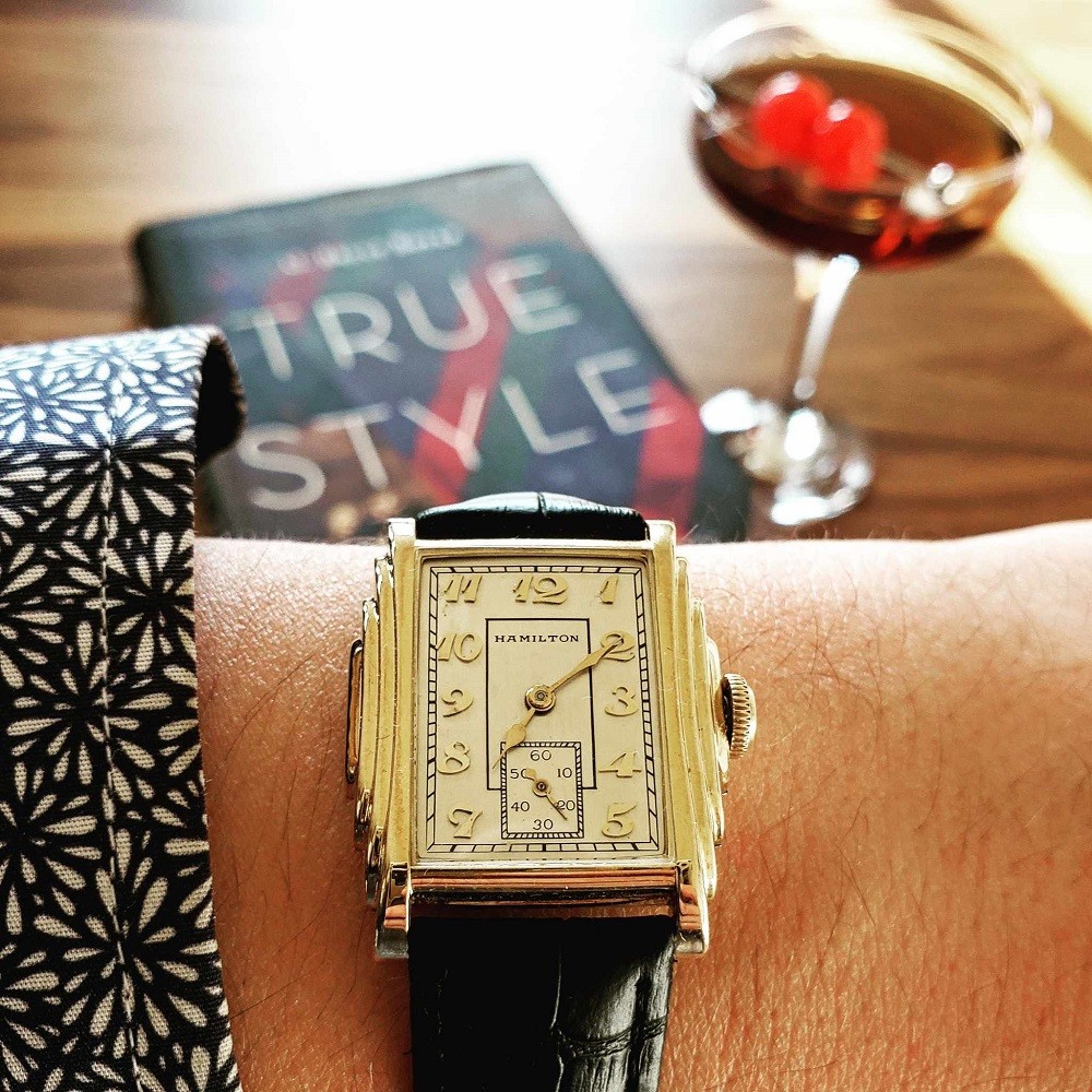 A vintage gold Hamilton watch features a white face, gold hands, and a gold art-deco style stepped case. It has a black leather strap