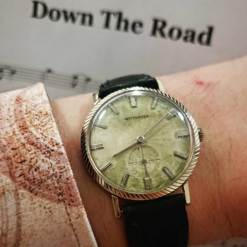 A 1960s round Wittnauer watch has a green/brown patina dial. The case is silver and features a knot-like design. The watch is on a black leather strap