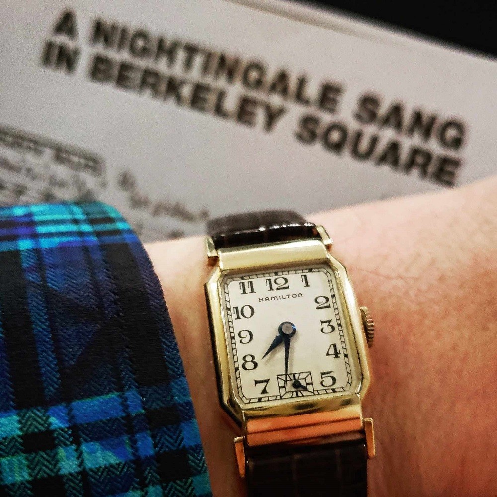 A vintage, gold, square Hamilton watch has black numerals, blued hands, and features a gold case. The watch is on a dark brown leather strap
