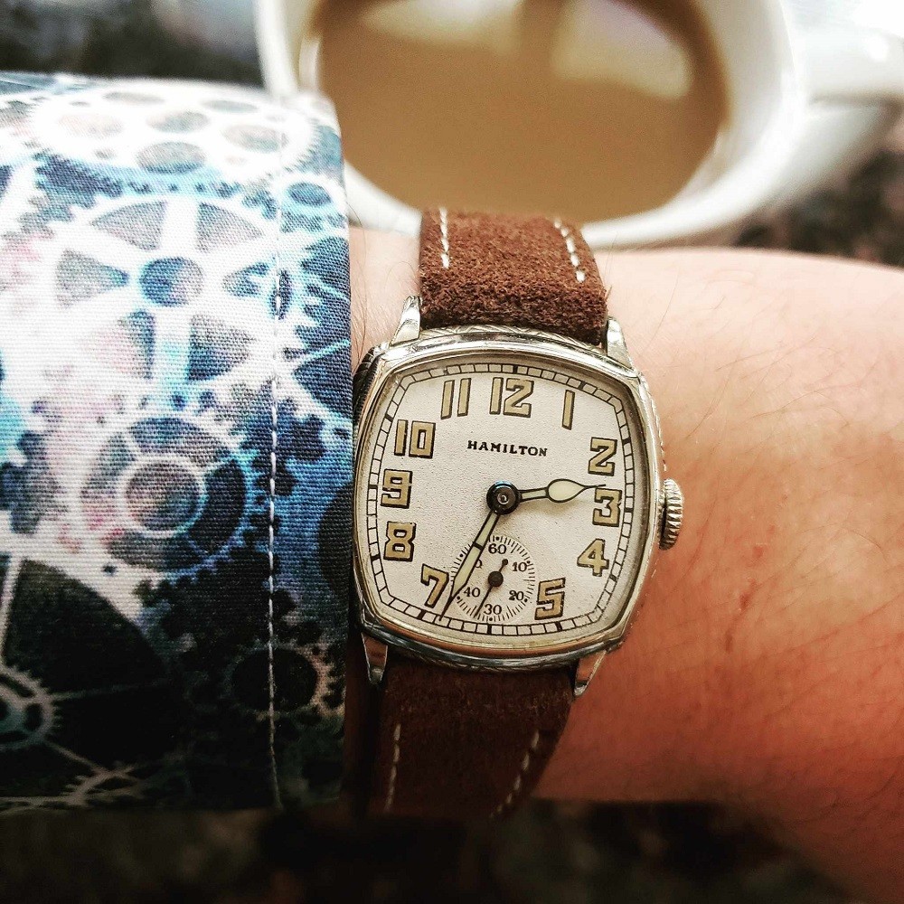 A vintage Hamilton watch in a silver square case with rounded edges has a brown suede strap. The numbers on the watch are yellow and the face of the watch is white