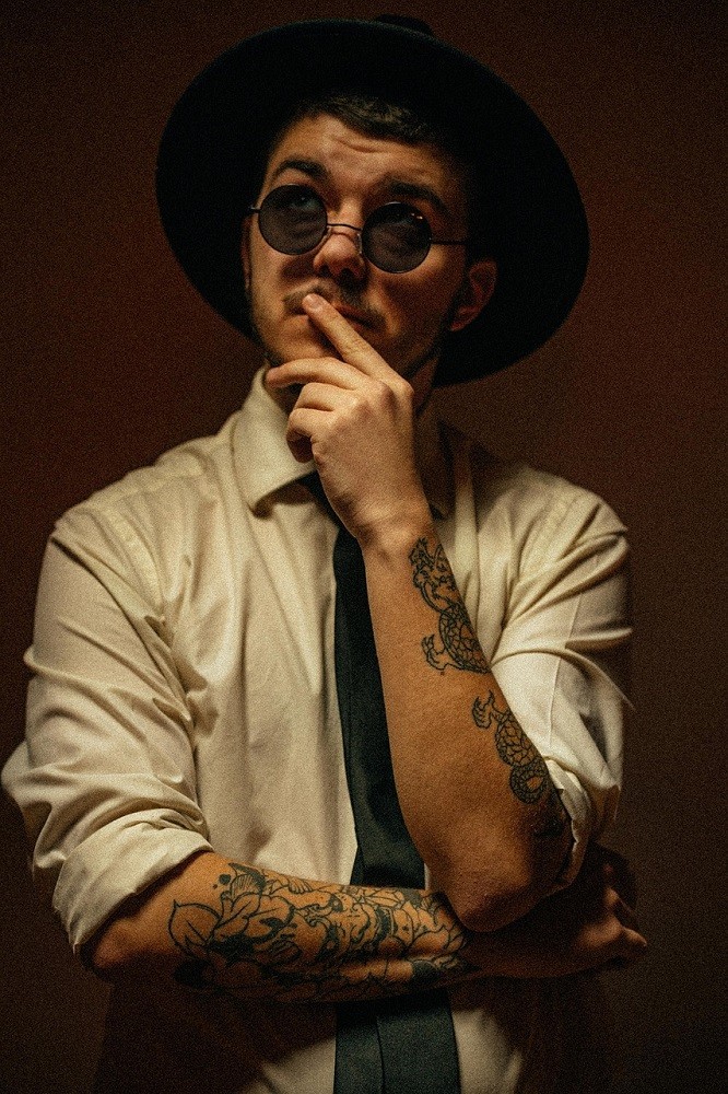 A man in a white dress shirt and black tie is wearing trendy round sunglasses and a black fedora