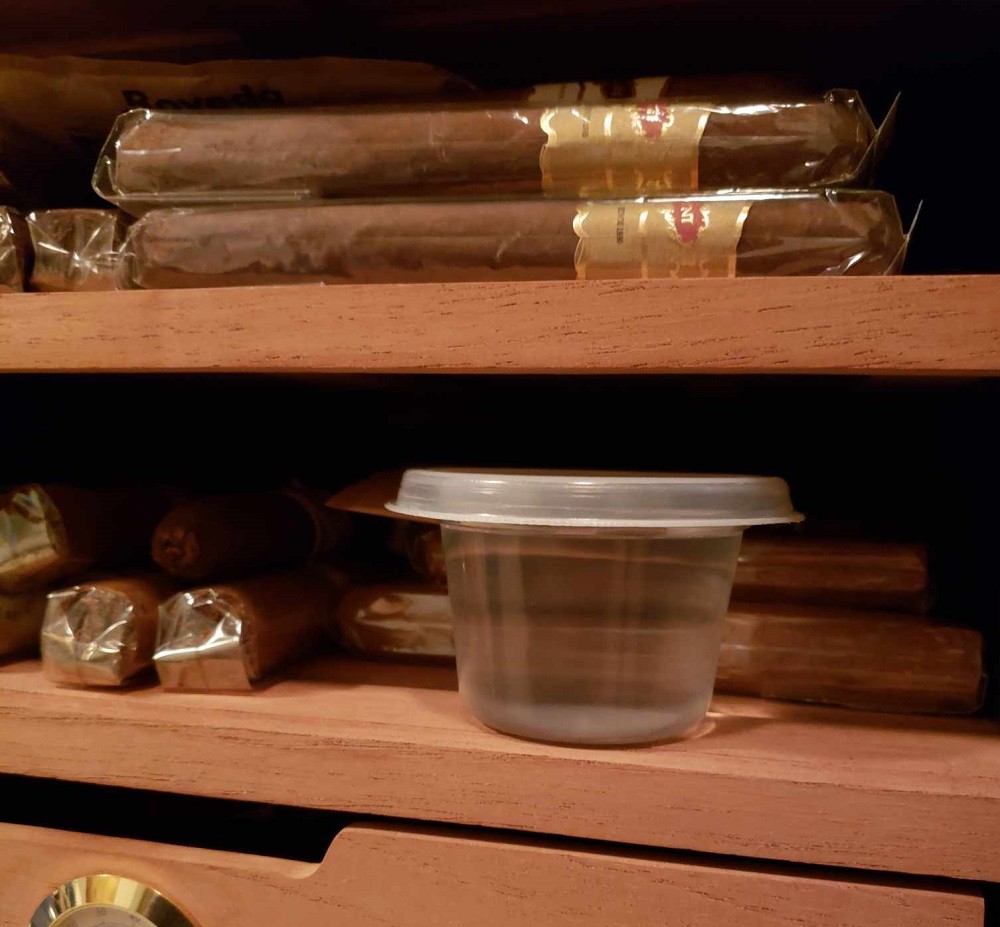 A small clear plastic cup of distilled water sits on a cedar shelf inside a cigar humidor