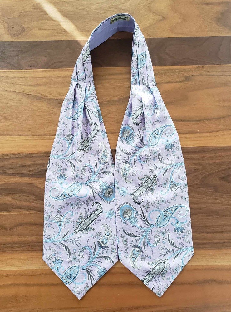 A pale pink ascot with a light blue and purple paisley design is laid out on a wooden table