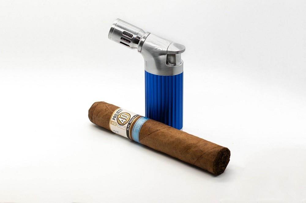 A light brown cigar with a white and teal blue band sits in front of a torch lighter. The lighter has a blue base and a silver top