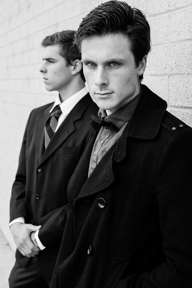 Two stylish men pose for the camera. They are wearing suits, ties, and dress shits. One man is staring directly at the camera and the other is looking away