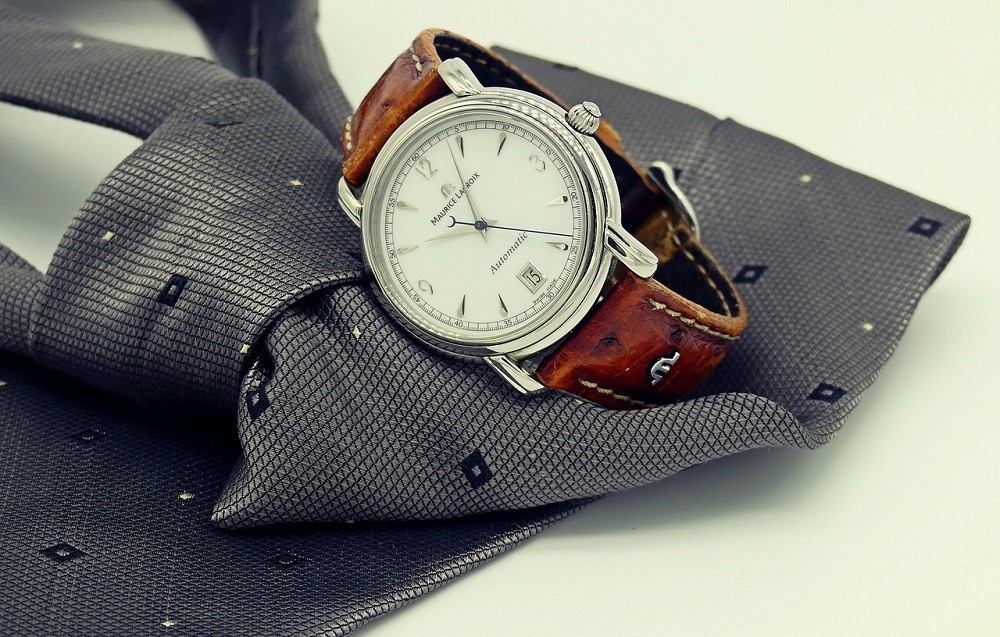 An expensive luxury watch with a white face and brown strap sits on a grey necktie