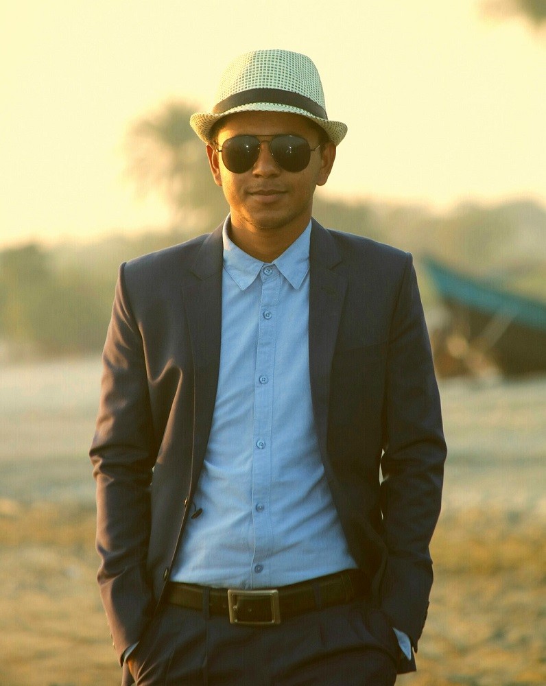 A man with darker skin is posing in front of the camera. He is wearing a white fedora hat, aviator sunglasses, a dark suit, and a light blue shirt