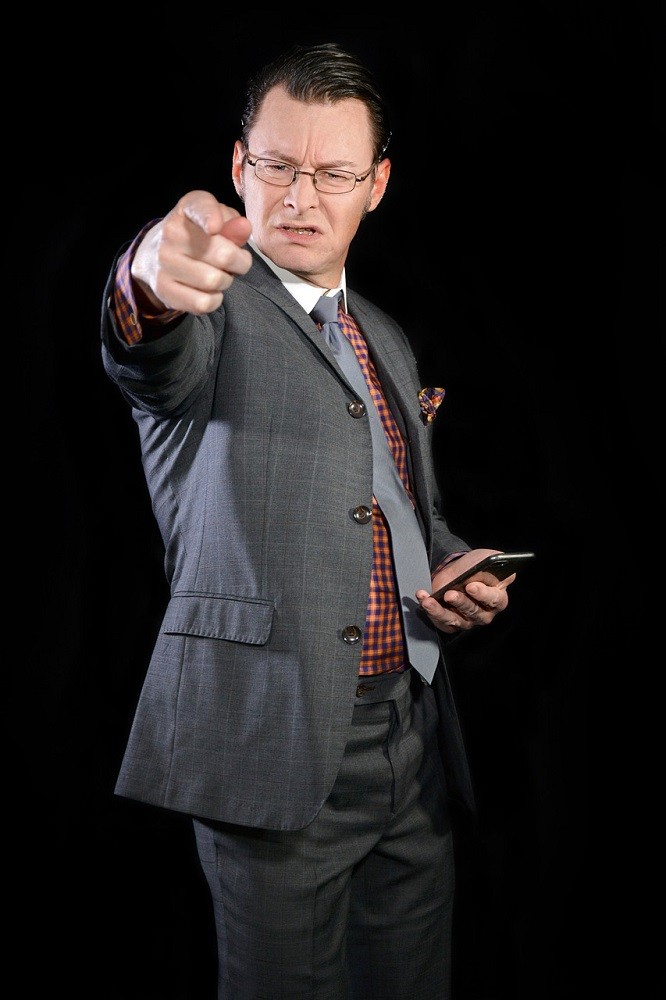 A man in a grey suit is pointing directly at the camera. He has a disgusted look on his face