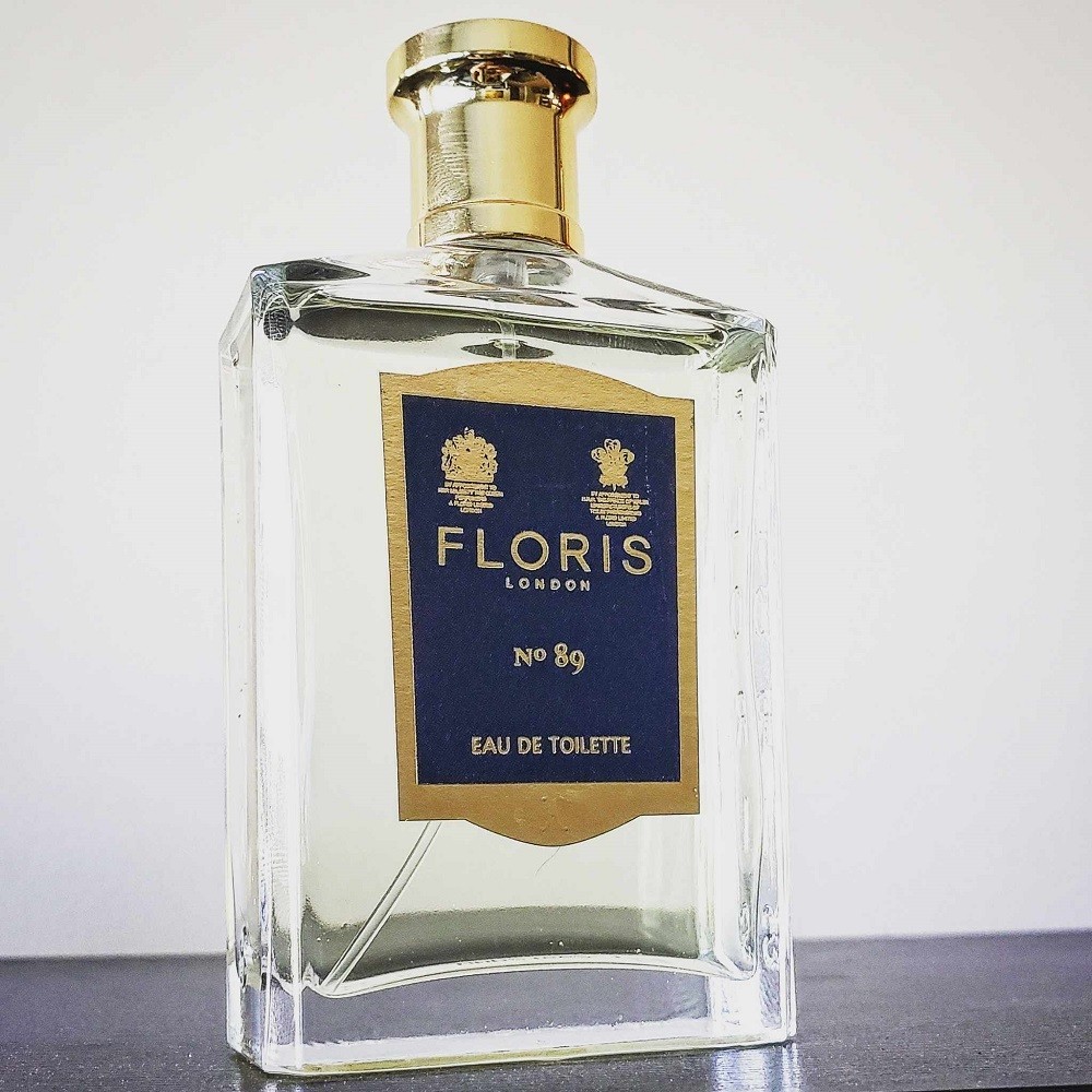 A clear rectangular bottle of No. 89 by Floris is on a black table. The cap is gold and the sticker on the front is dark blue with a gold border. The name of the fragrance and brand is in the label