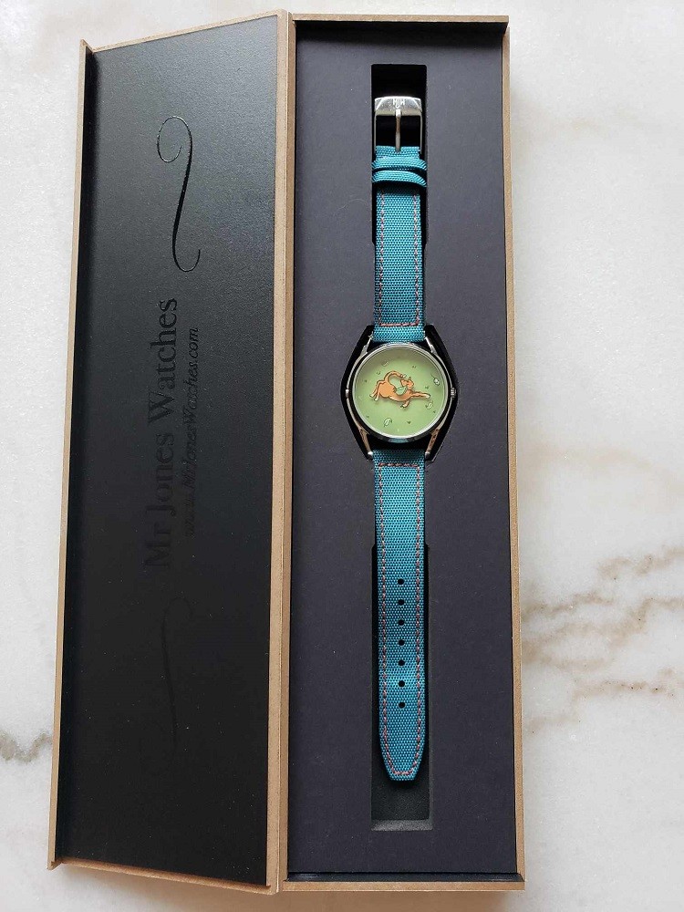The Mispoes watch by Mr Jones Watches features an orange cat with an outstretched paw on the dial. The strap is teal and it is sitting inside the presentation box