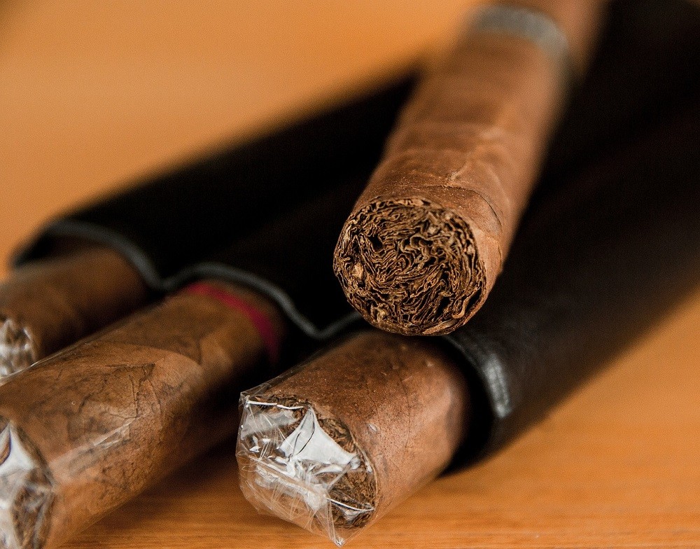 Three brown cigars lay flat on a table. Two are in a black leather travel case and one is laying on top