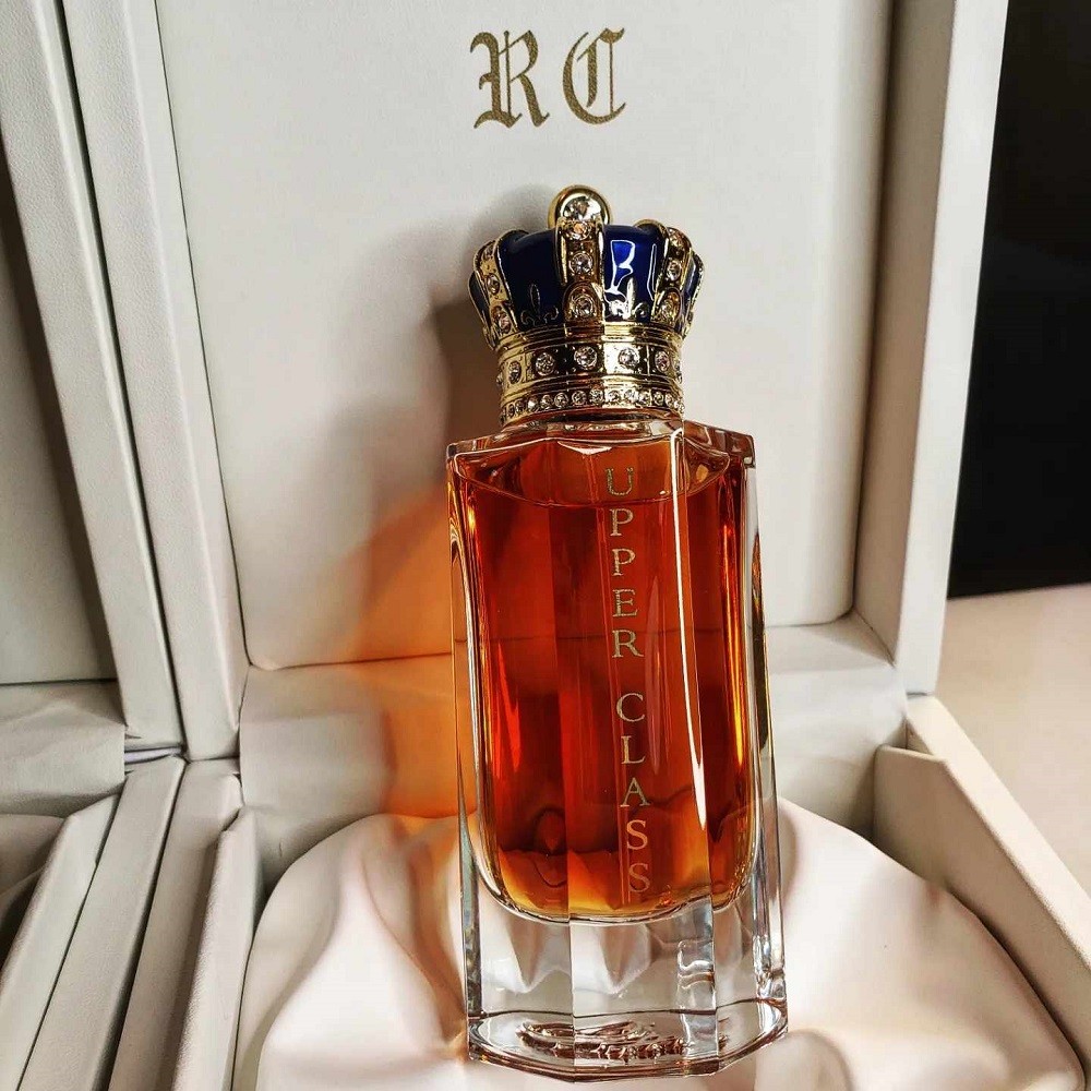 A bottle of Upper Class by Royal Crown luxury cologne sits perched up inside its white display box. The bottle is a tall glass rectangle with thick glass. It has a blue and gold decorative crown as the cap