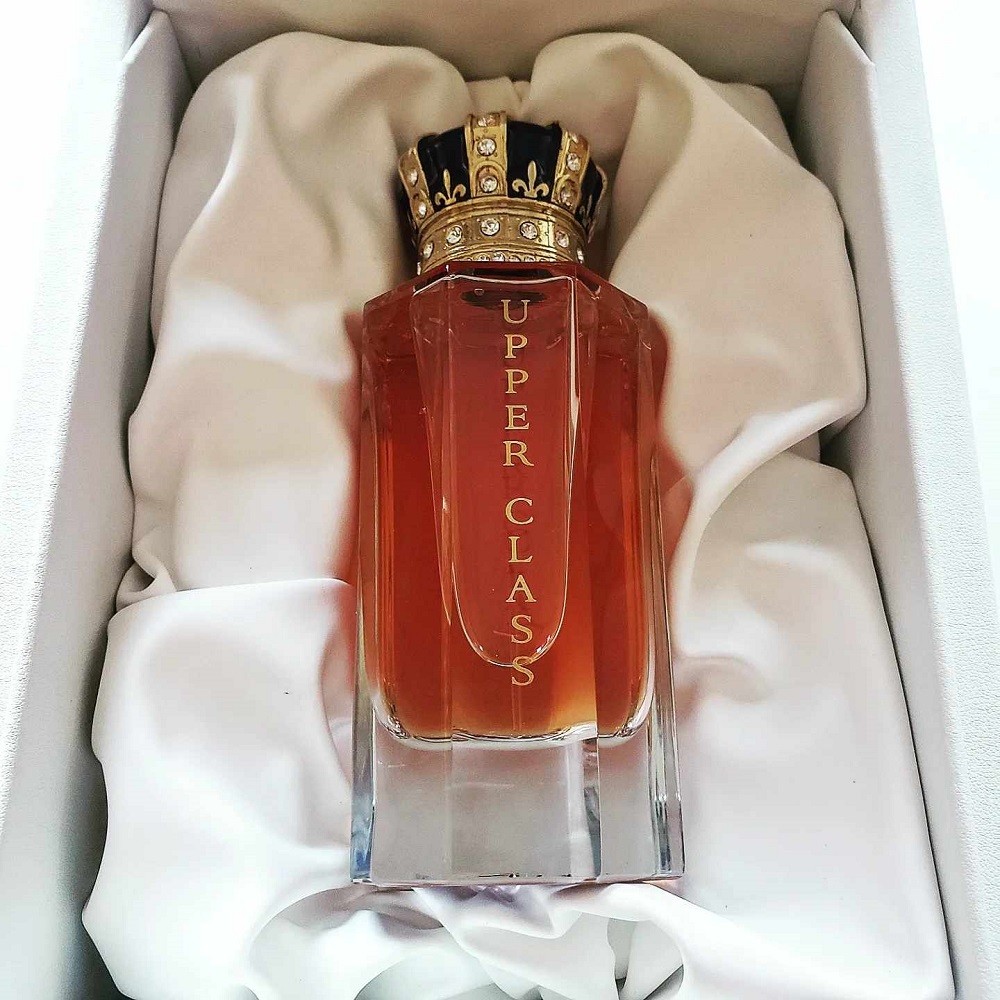 A bottle of light brown cologne with a blue and gold crown sits in a satin sheet inside its display box
