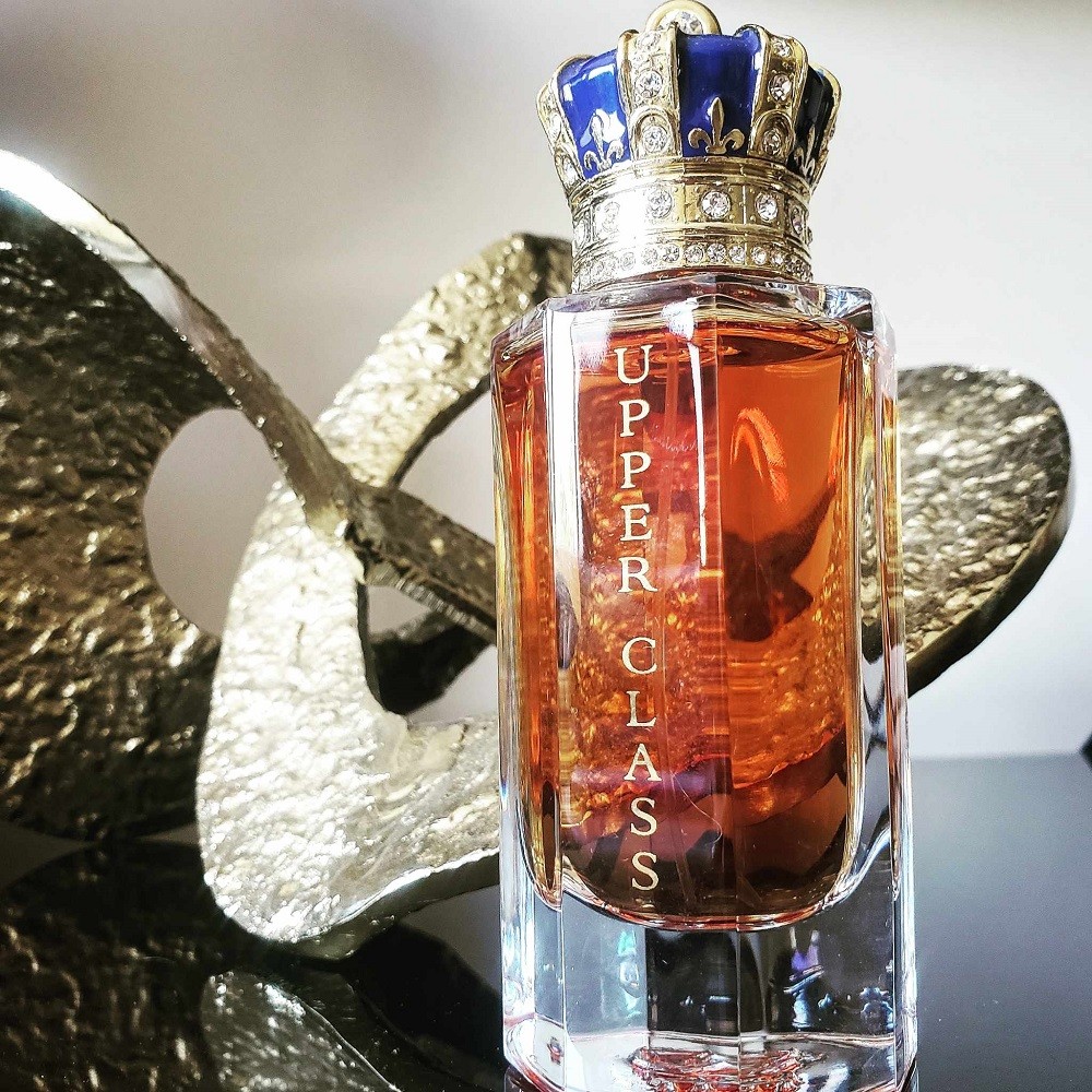 A thick glass fragrance bottle has a blue and gold crown cap. It sits beside a gold piece of abstract art