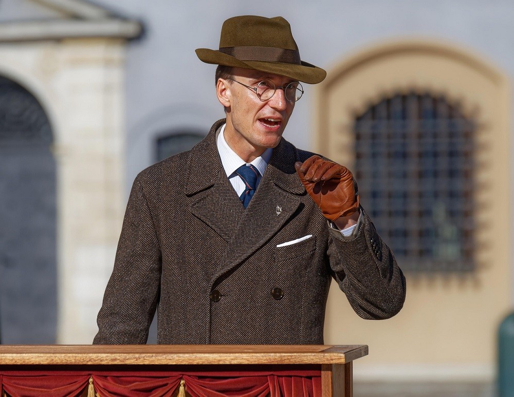 A man wearing a brown wool suit with a white pocket square and matching brown fedora is standing at a podium giving a speech