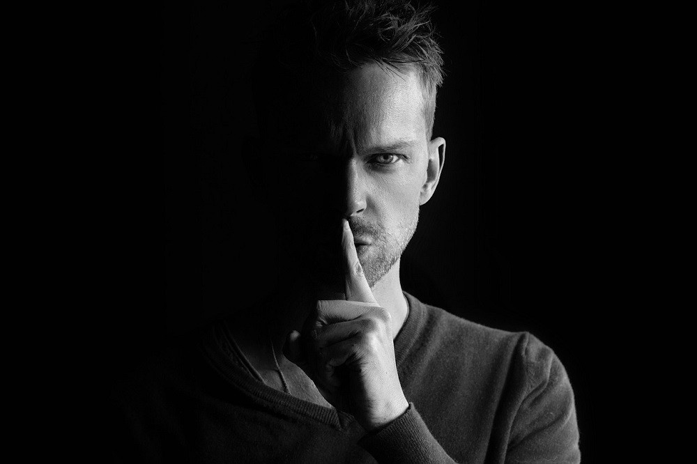 A black and white photograph of a younger man with his index finger extended over his lips making a 