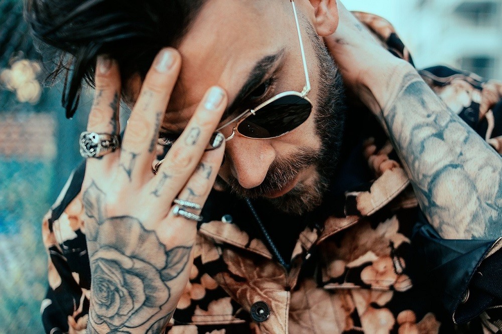A man wearing aviator sunglasses, multiple chunky rings, and sporting a large beard has his hand on his head as in disbelief of whatever he just saw or heard