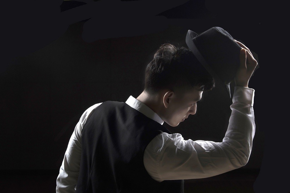 A young man wearing a white shirt and black vest tips his black fedora to someone outside of the picture