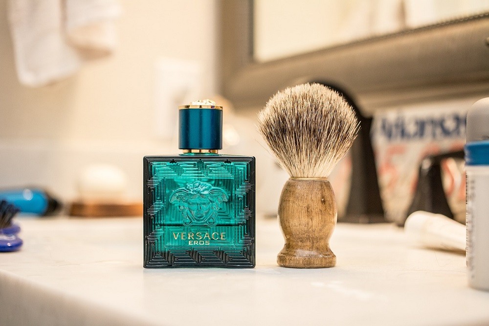 A rectangular, teal bottle of Eros by Versace cologne sits on a bathroom counter with a shaving brush beside it. The shaving brush has a light coloured wooden handle and dark bristles