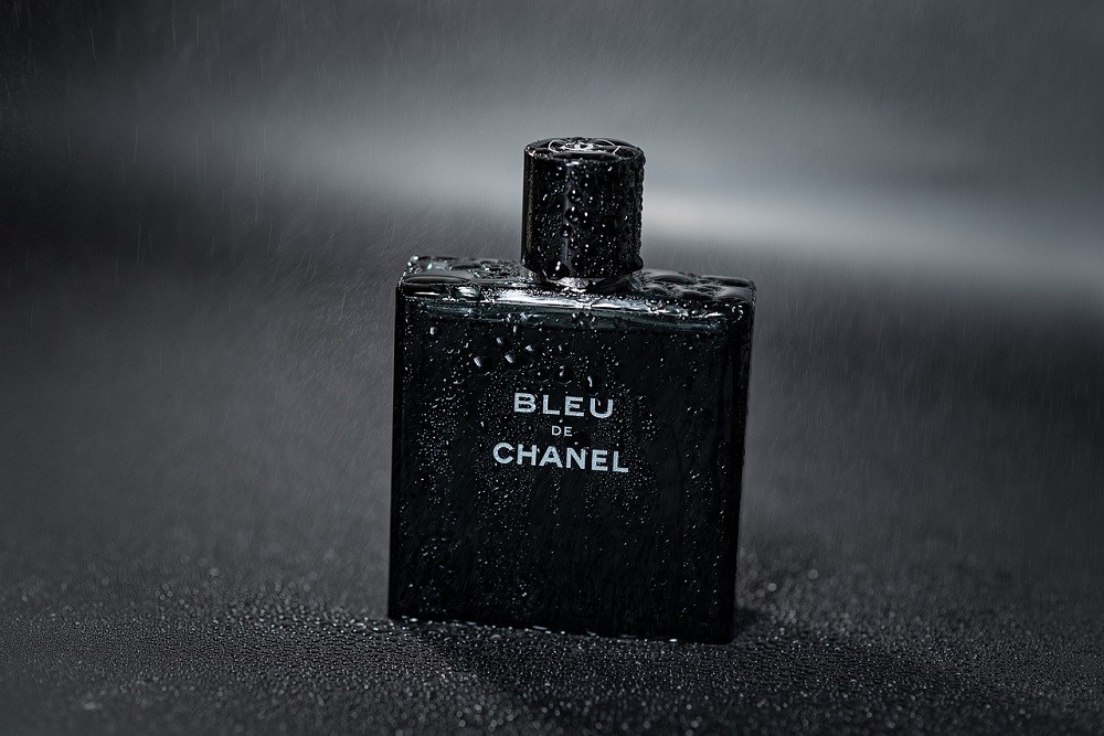 A blue/black square bottle of Bleu de Chanel cologne is sitting in black sand. The bottle has condensation on it