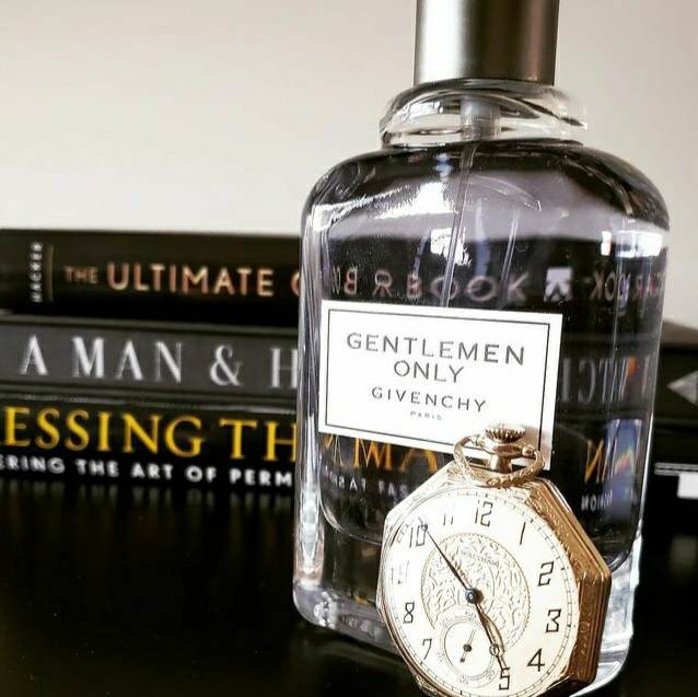 A clear bottle of Gentleman Only cologne by Givenchy sits beside some men's style books. An octagonal vintage pocket watch rests against the bottle