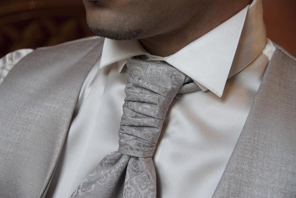 A formal grey ascot with paisley pattern is tied on top of a plain white shirt. The man in the picture is wearing a grey vest