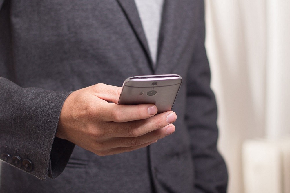 A picture of a man from the neck down. He's wearing a grey sport coat and whtie shirt. He has his smartphone in his hand facing away from the camera