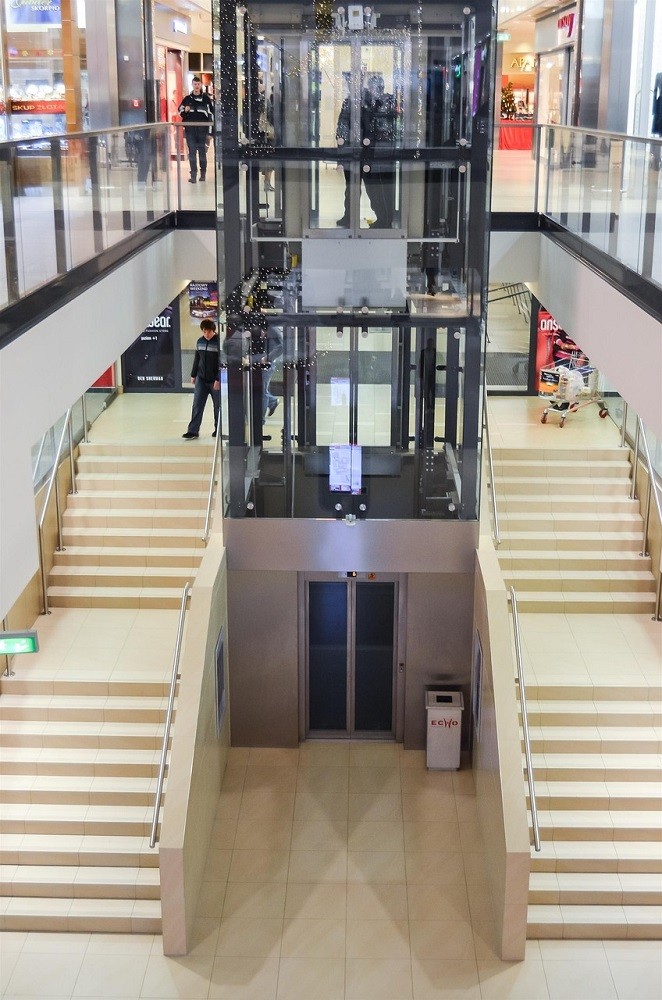 A glass elevator is at a mall. There are stairs on each side of the elevator