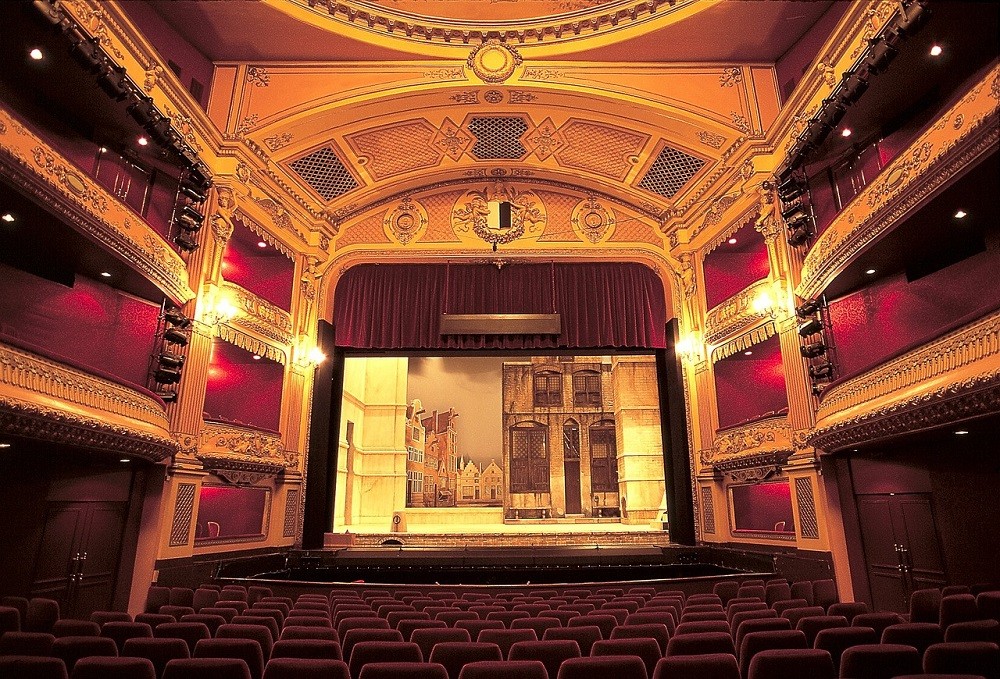 A view of the theatre stage from the back of the main seating area. There are two balconies on each side. The seats are red velvet, the wood is painted gold, and there's a big red curtain above the stage