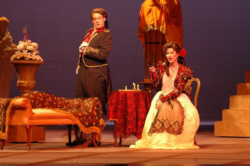 A male and female performer are on stage dressed in fancy Victorian era clothes. The set is filled with a small table with a red velvet cloth, an orange bench, and a large vase with white and purple flowers in it