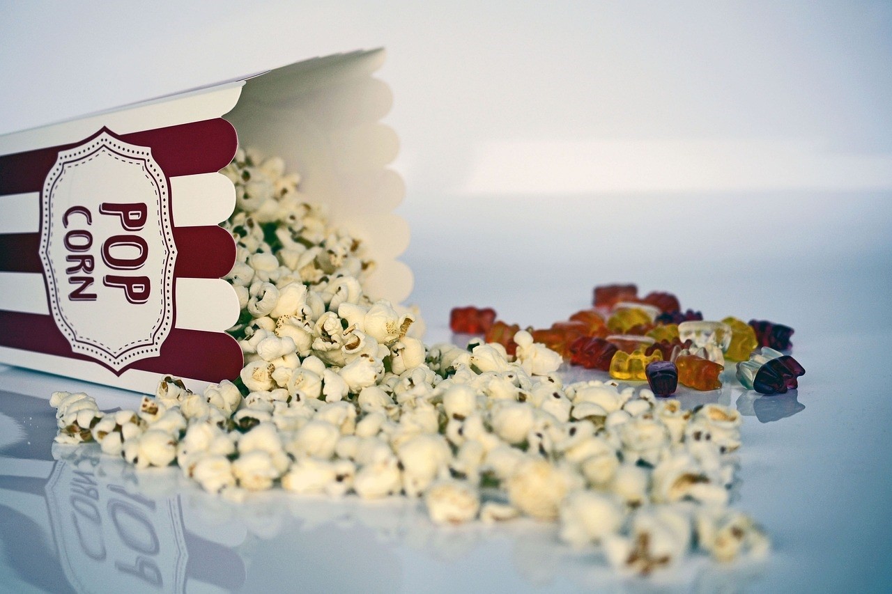A red and white box of popcorn is on its side. Popcorn has spilled out. Behind the popcorn is a handful of gummy bears