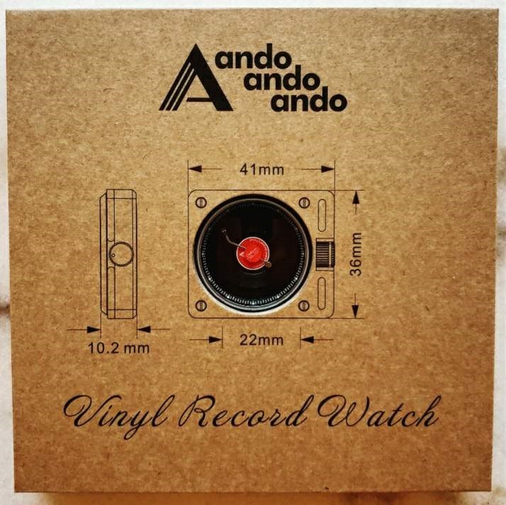 The front of the box that contains the Vinyl Record Watch by AndoAndoAndo. The name of the brand and brand logo is at the top, a display window features the dial of the watch including the measurements for the case. The name of the watch is at the bottom in a cursive font