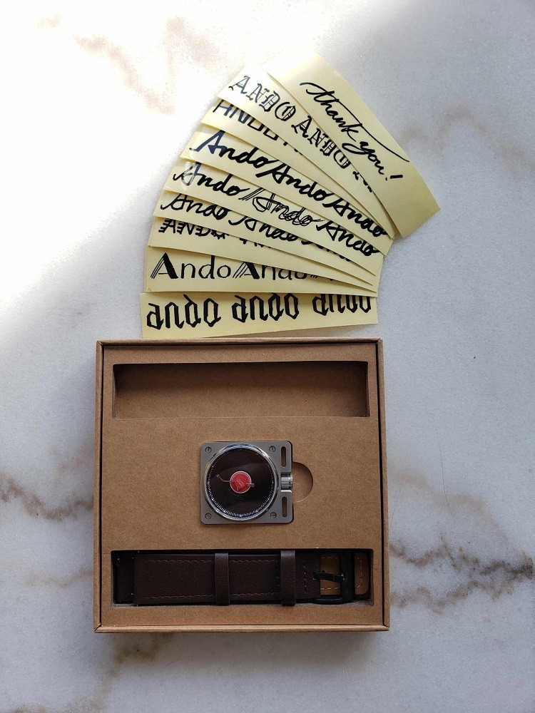 A front view of the box that contains the Vinyl Record Watch by AndoAndoAndo including the several stickers with the brand name in a variety of fonts. The box and strap sit inside the thick cardboard box