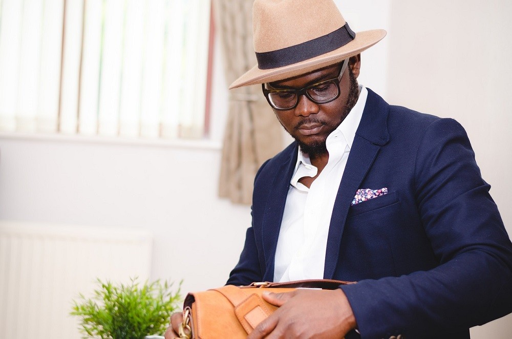 A man wearing a beige fedora sports a dark blue sport coat and white dress shirt. He has a lively pocket square with a small repeating pattern in his breast pocket