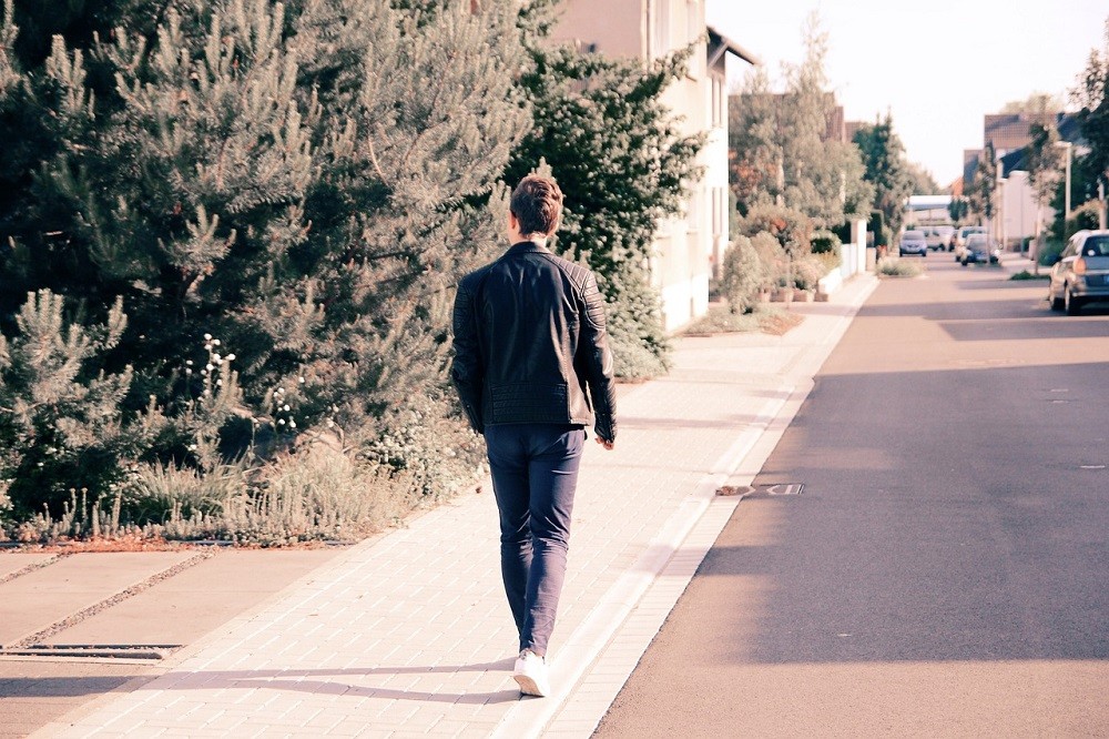 A man walks on a sidewalk walking against traffic. This is the safest way to walk on a sidewalk because you can see oncoming traffic
