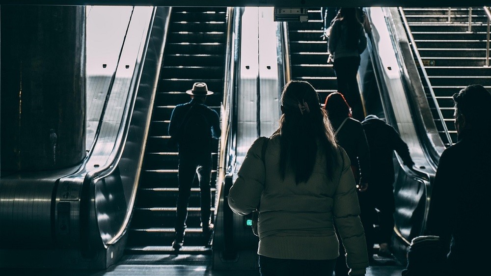People board an escalator going up. They are spaced apart and all getting on on the right side, leaving space on the left for people to walk up