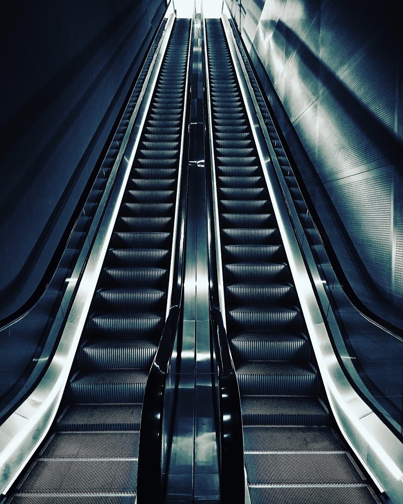 Two escalators are side-by-side. They are in a dimly lit area, possibly leading to a subway station or underground train. No one is riding them at the moment