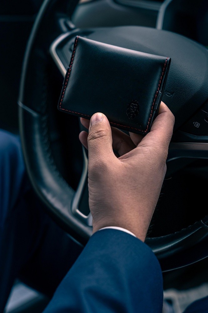 A man holds a black wallet in his hands. He is sitting in the driver seat of his car. The steering wheel is in the background