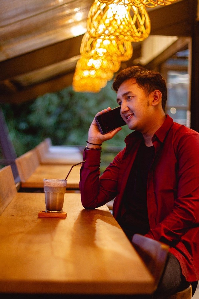 A young man dressed in a red jacket sits at a table at a restaurant. He is on his phone while he waits for his food to be delivered