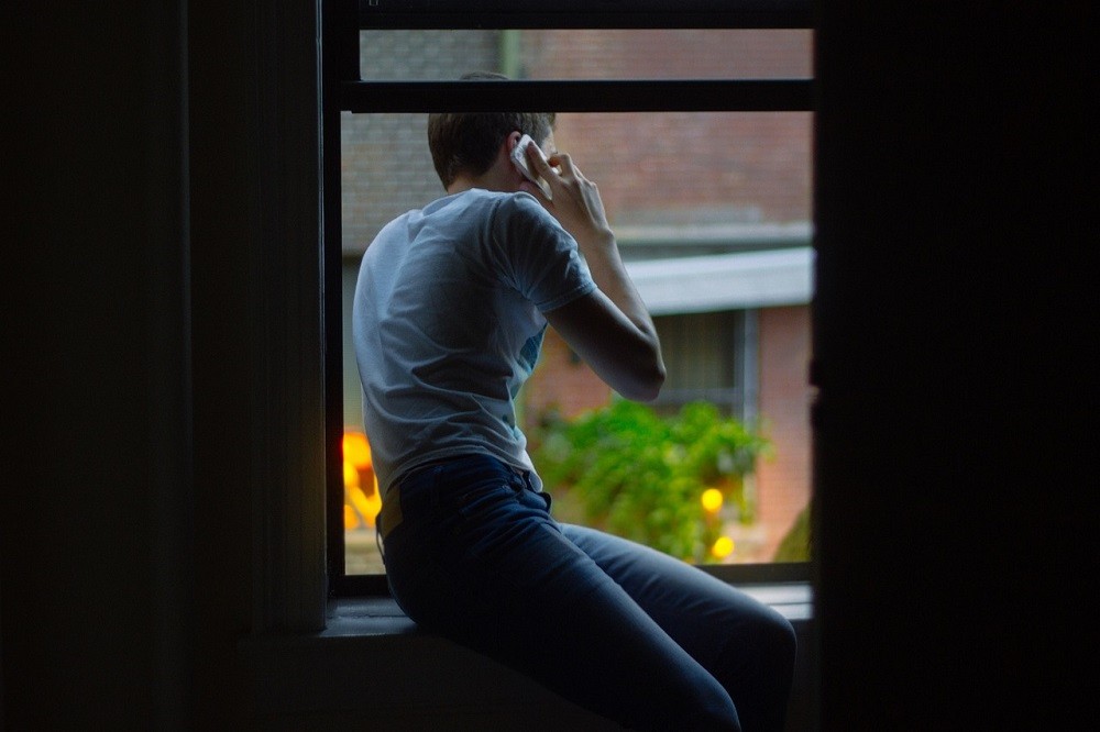 A young man sits on his windowsill while on the phone. He is wearing jeans and a grey t-shirt