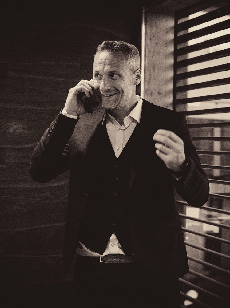 A black and white picture of a businessman on his cell phone. He is grinning with excitement with his arms raised by his shoulders