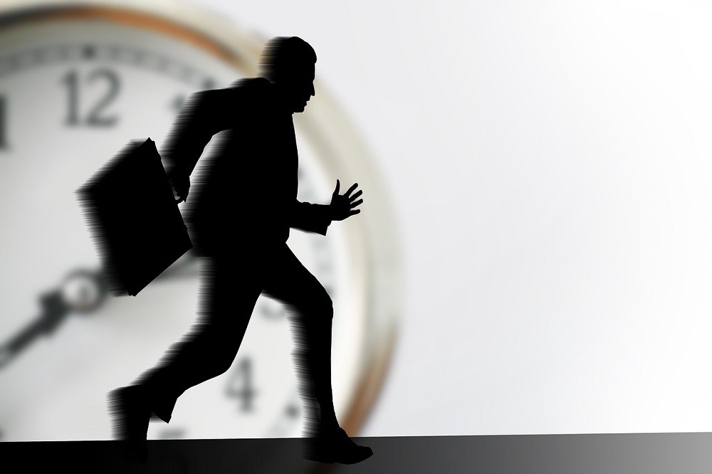 A shadow of a businessman with a briefcase is running so fast he is becoming blurry. There is a large clock in the background, signifying the importance of time