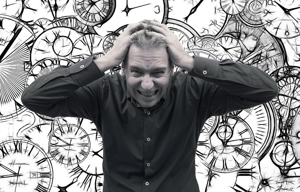 A man is visibly stressed out. He has his hands on his head. In the background are several clocks showing different times