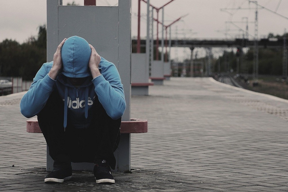 A young man in a blue hoodie and black pants sits alone along a train station with his head in his hands. He looks scared