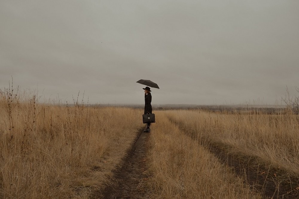 A person dressed in all black stands alone in a field with an overcast sky