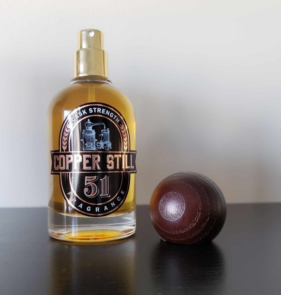 The bottle of 51 by Copper Still Fragrance is a dark amber colour. The bottle sits beside the dark brown, round, wooden cap