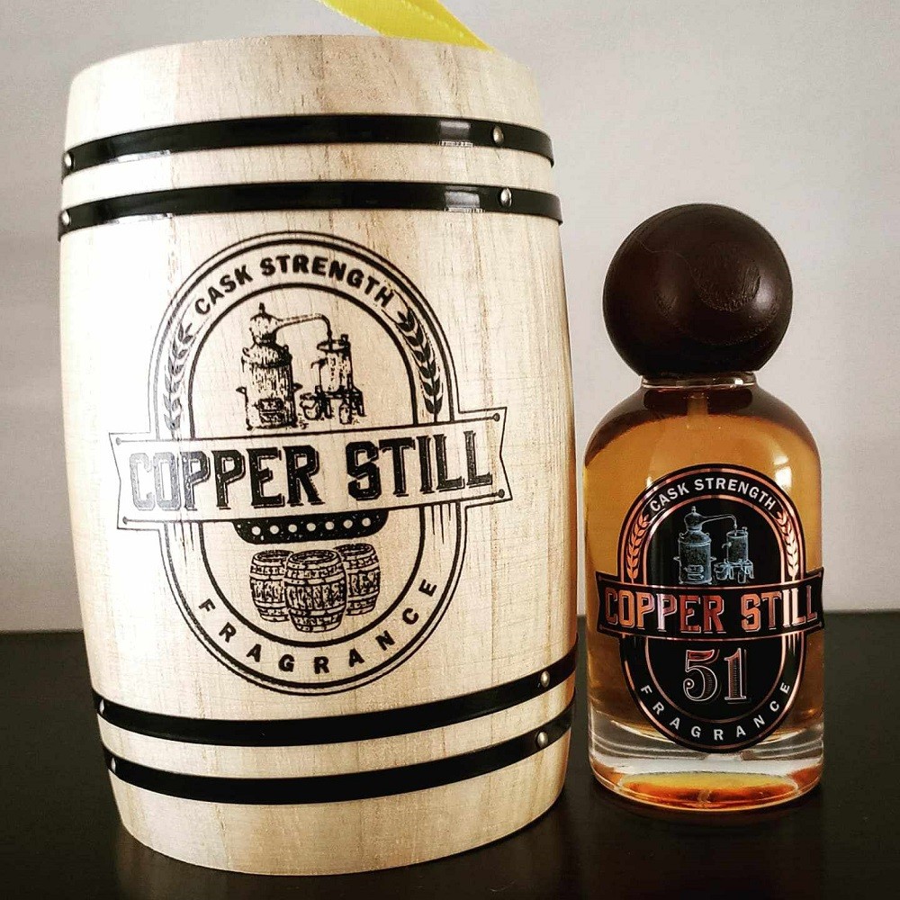 A clear glass bottle of Copper Still Fragrance 51 fragrance sits beside the miniature oak barrel box it comes in